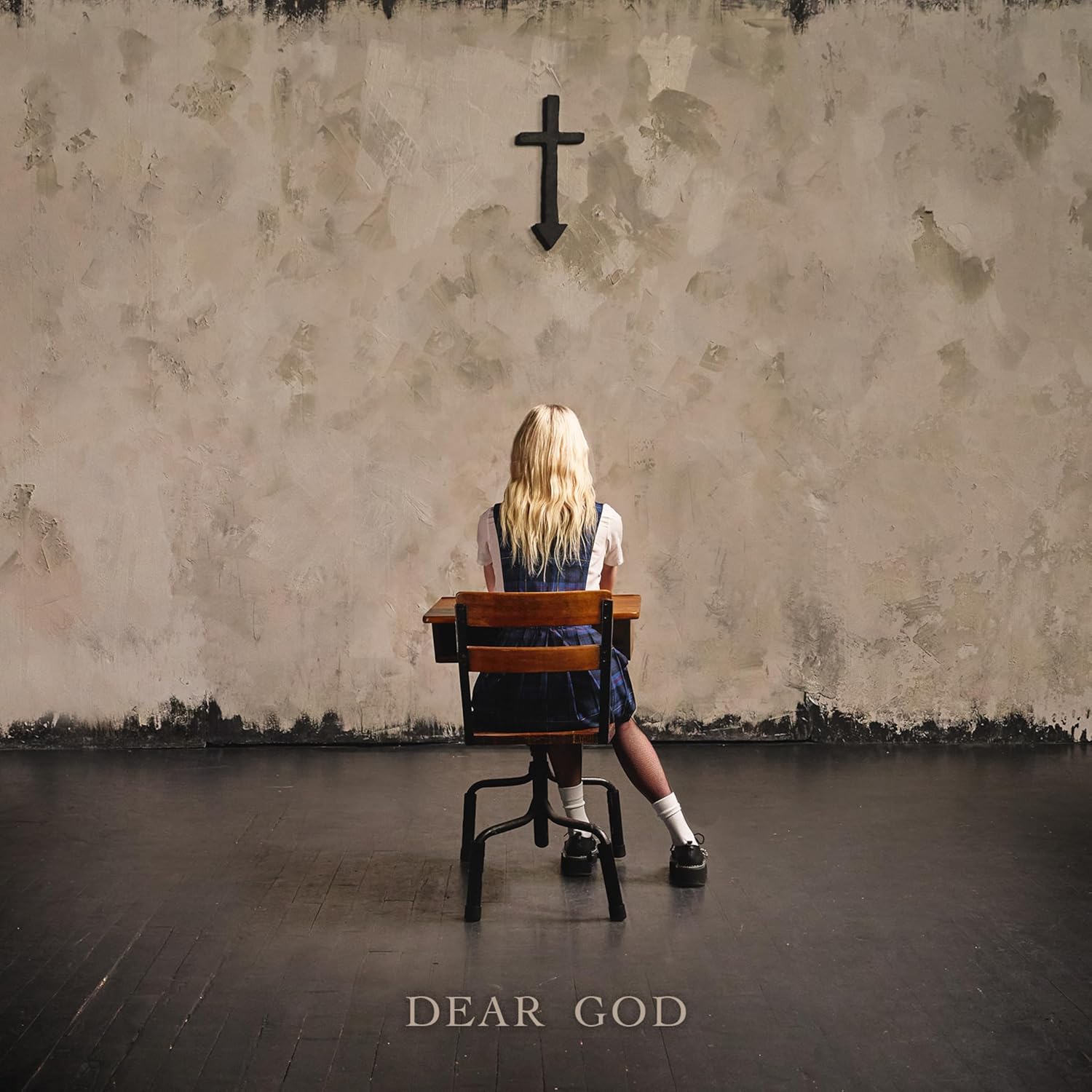 A blonde girl sits at a school desk facing a wall with an inverted cross.