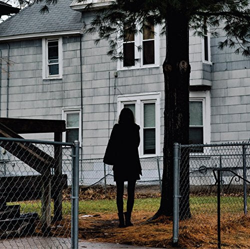 A woman in black stands before a fenced grey house with a tree.