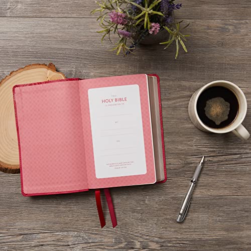 Open pink Bible with a presentation page, coffee mug, and pen on wooden table.
