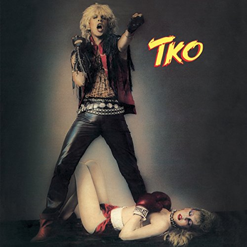 A blonde man in leather stands over a woman wearing boxing gloves on the floor.
