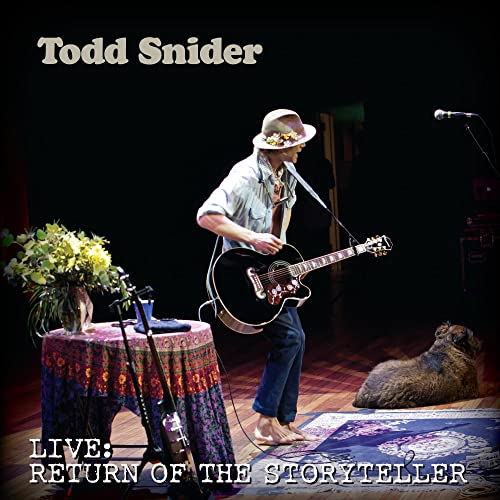 Todd Snider playing acoustic guitar barefoot on stage with a dog resting nearby.