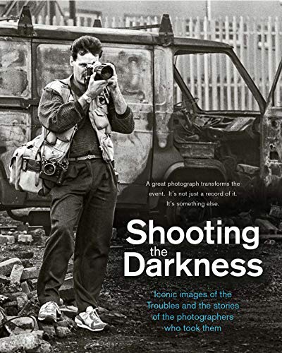 A black and white photo of a photographer standing before a damaged van.