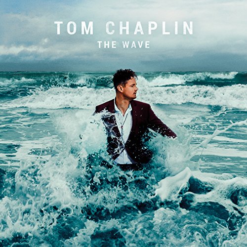 Man in a suit standing in ocean waves under cloudy sky.
