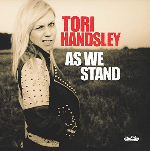 The image depicts the album cover for "As We Stand" by Tori Handsley. To describe the image in 15 words or less for accessibility purposes, focusing on key visual characteristics and the main subject without marketing language, the alt text could be: "Blonde woman in black and white jacket standing in a field, album cover."