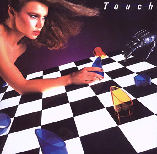 A woman reaches across a black and white checkered board toward a robotic hand.