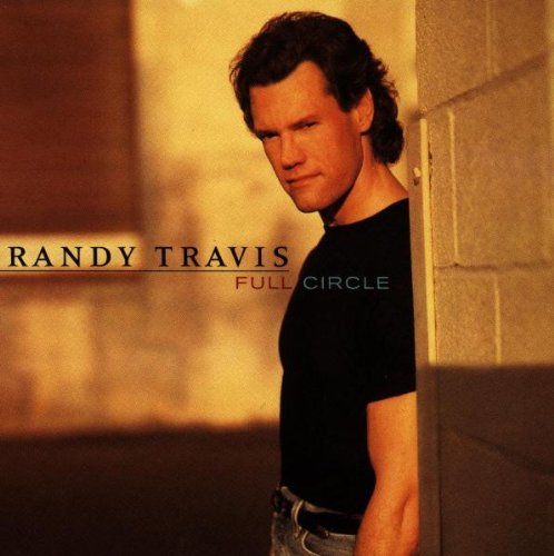 Randy Travis wearing a black t-shirt, leaning against a wall in warm lighting.