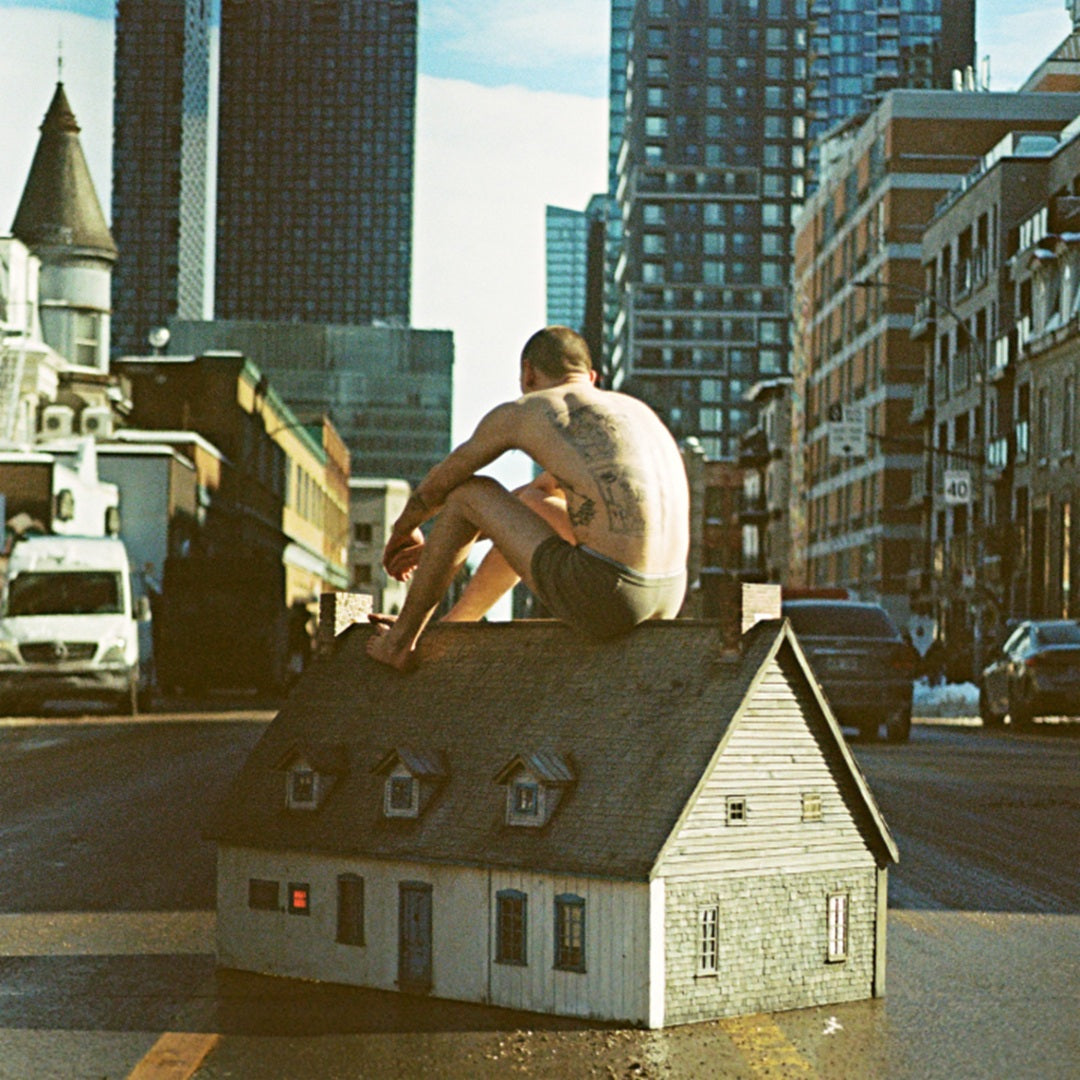 A shirtless tattooed man sits on the roof of a miniature house in a city.