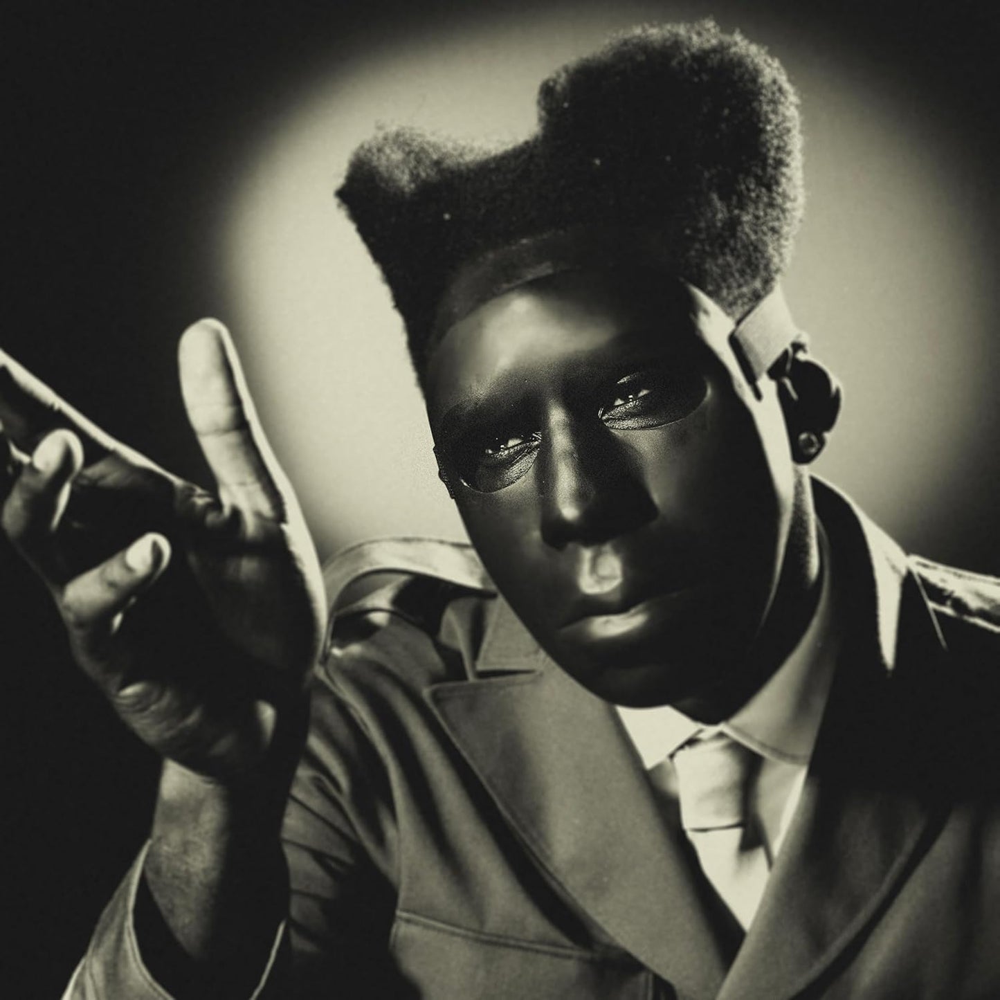 Tyler, the Creator wearing a black mask and suit, gesturing with his hand.