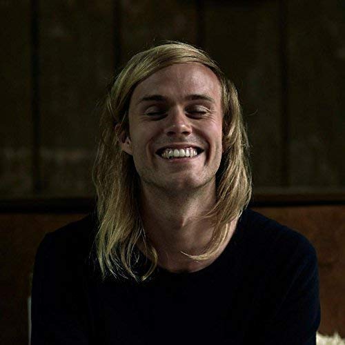 A smiling person with long blonde hair against a dark background.