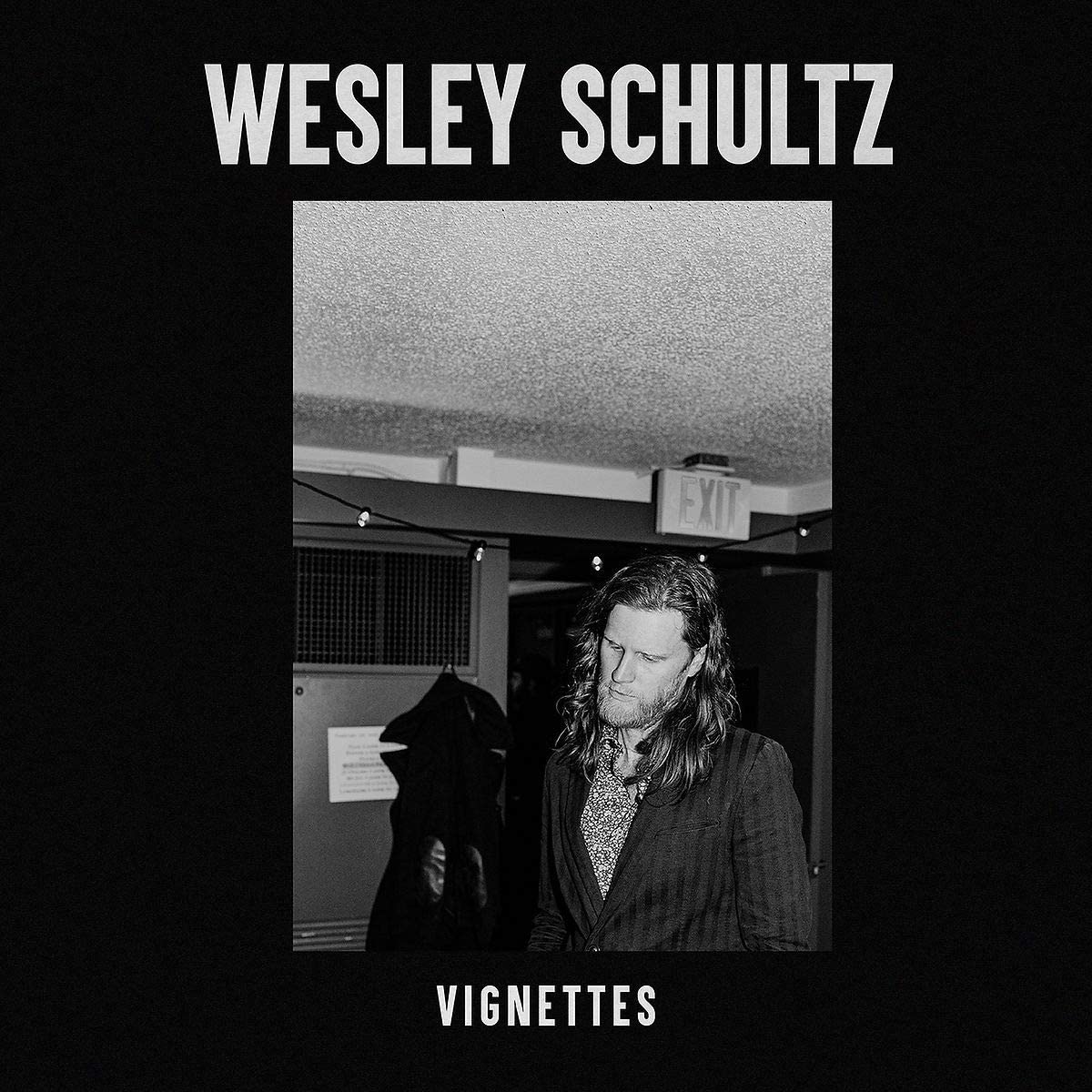 The image is an album cover for "Vignettes" by Wesley Schultz. The alt text is: "Black and white album cover with a man and exit sign."