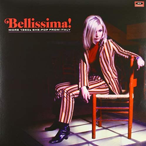 A blonde woman in a striped suit sits on a red chair on a tiled floor.