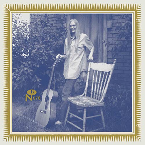 A person with long hair stands holding an acoustic guitar next to a wooden chair.