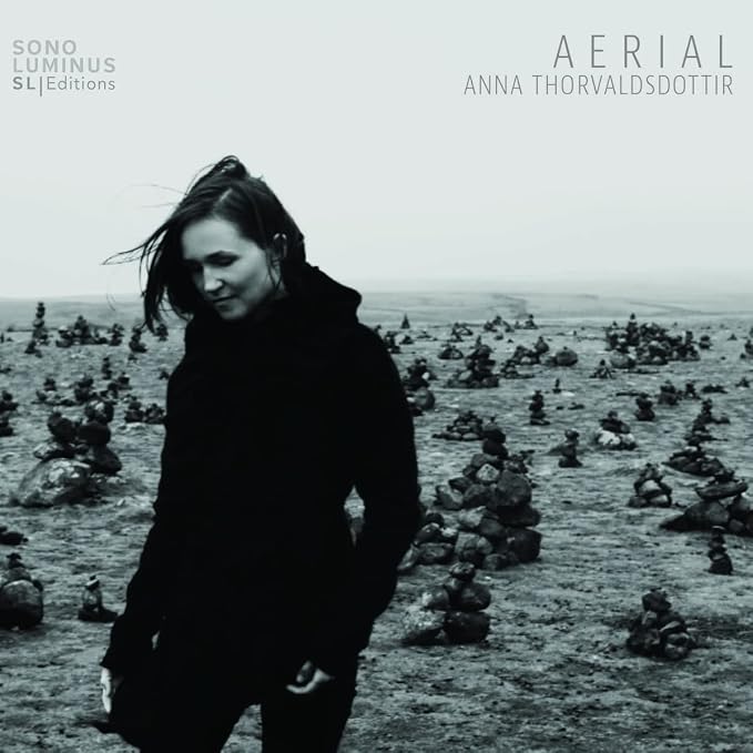 CD cover: Woman in black coat amidst stone cairns, grayscale.