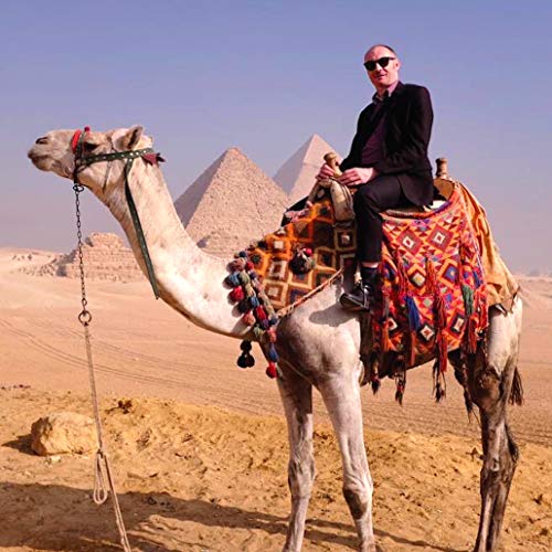 Man in suit riding a camel with pyramids in the background.