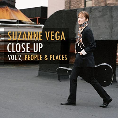 Suzanne Vega walking with a guitar case in a city street.