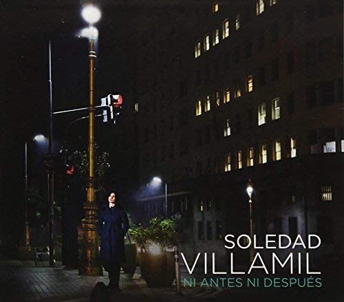 Woman in dark coat standing on city street corner at night with streetlights.
