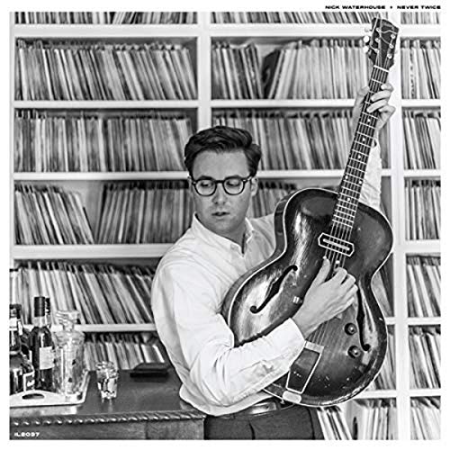 Man in glasses and white shirt playing an acoustic guitar in front of bookshelves.