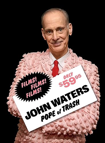 John Waters in pink fluffy coat, holding a price-tagged sign.