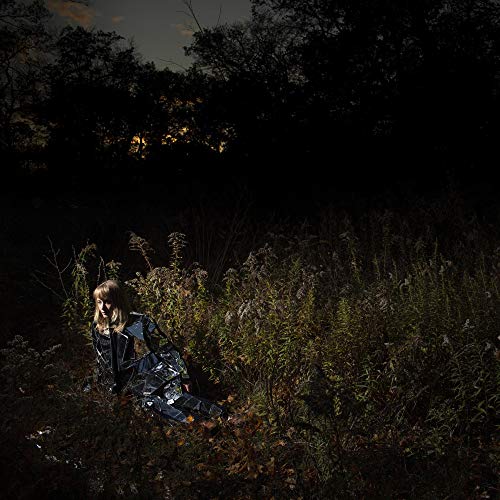 A woman sits in tall grass and weeds, surrounded by dark trees and foliage.