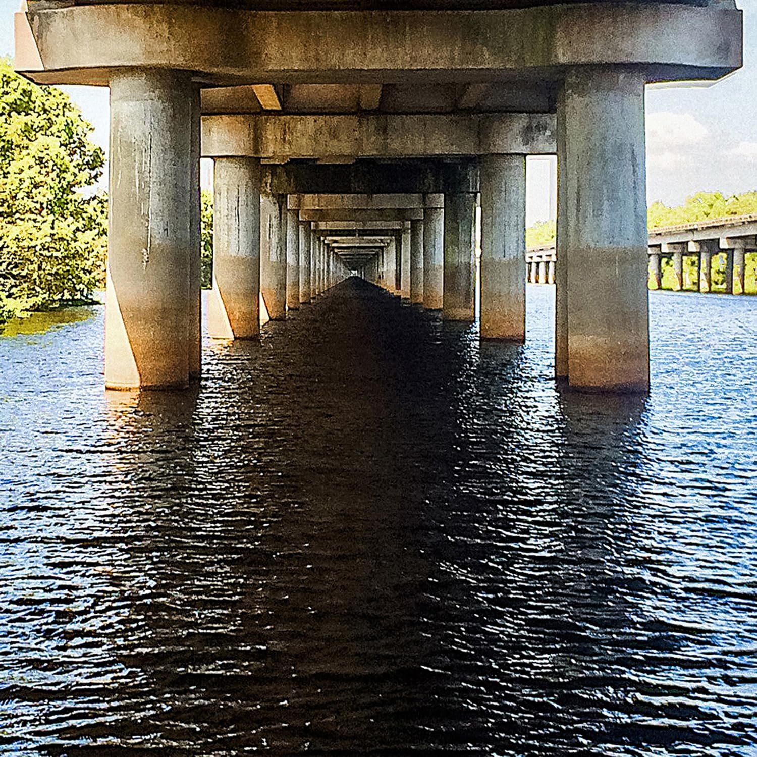 Symmetrical view looking beneath a long concrete bridge over dark rippling water.