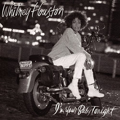 Whitney Houston sitting on a motorcycle with a city skyline background in black and white.