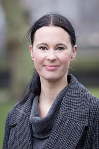 A woman with dark hair in a grey coat against a blurred outdoor background.