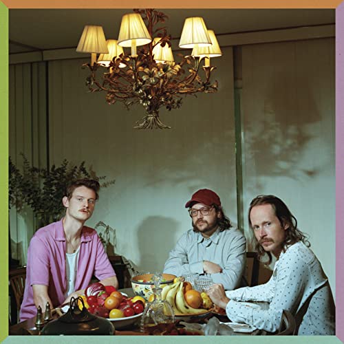 Three men sit at a dining table with a bowl of fruit under a chandelier.