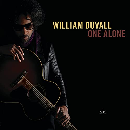William Duvall wearing sunglasses and holding an acoustic guitar against a black background.