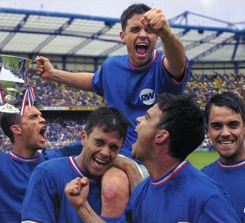 Robbie Williams on shoulders, celebrating with a trophy in a stadium.
