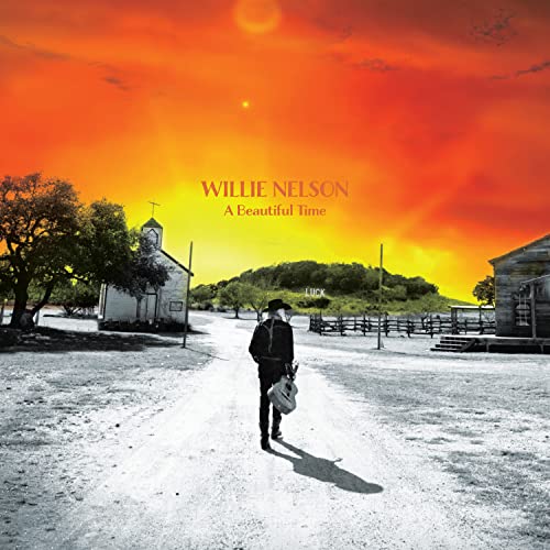 Willie Nelson walks down a dusty road toward buildings under a fiery sky.