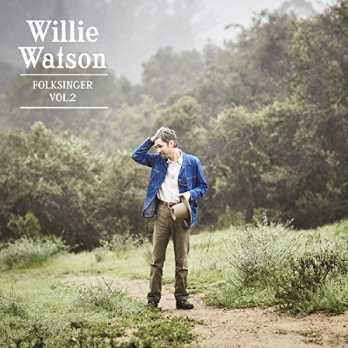 Man in blue jacket and khaki pants stands on a dirt path in a green landscape.