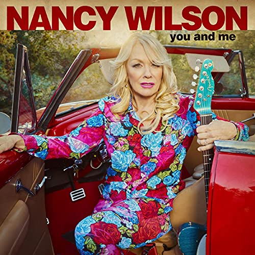 Nancy Wilson sits in a red convertible wearing a floral outfit, holding an electric guitar.