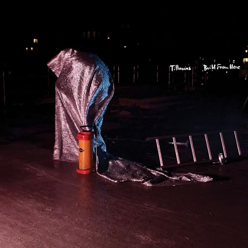 Figure draped in silver fabric stands in a dark, urban nighttime scene.
