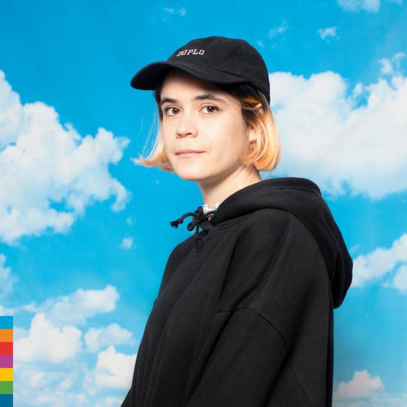 A person with short blonde-tipped hair wearing a black hoodie and Duplo cap against clouds.