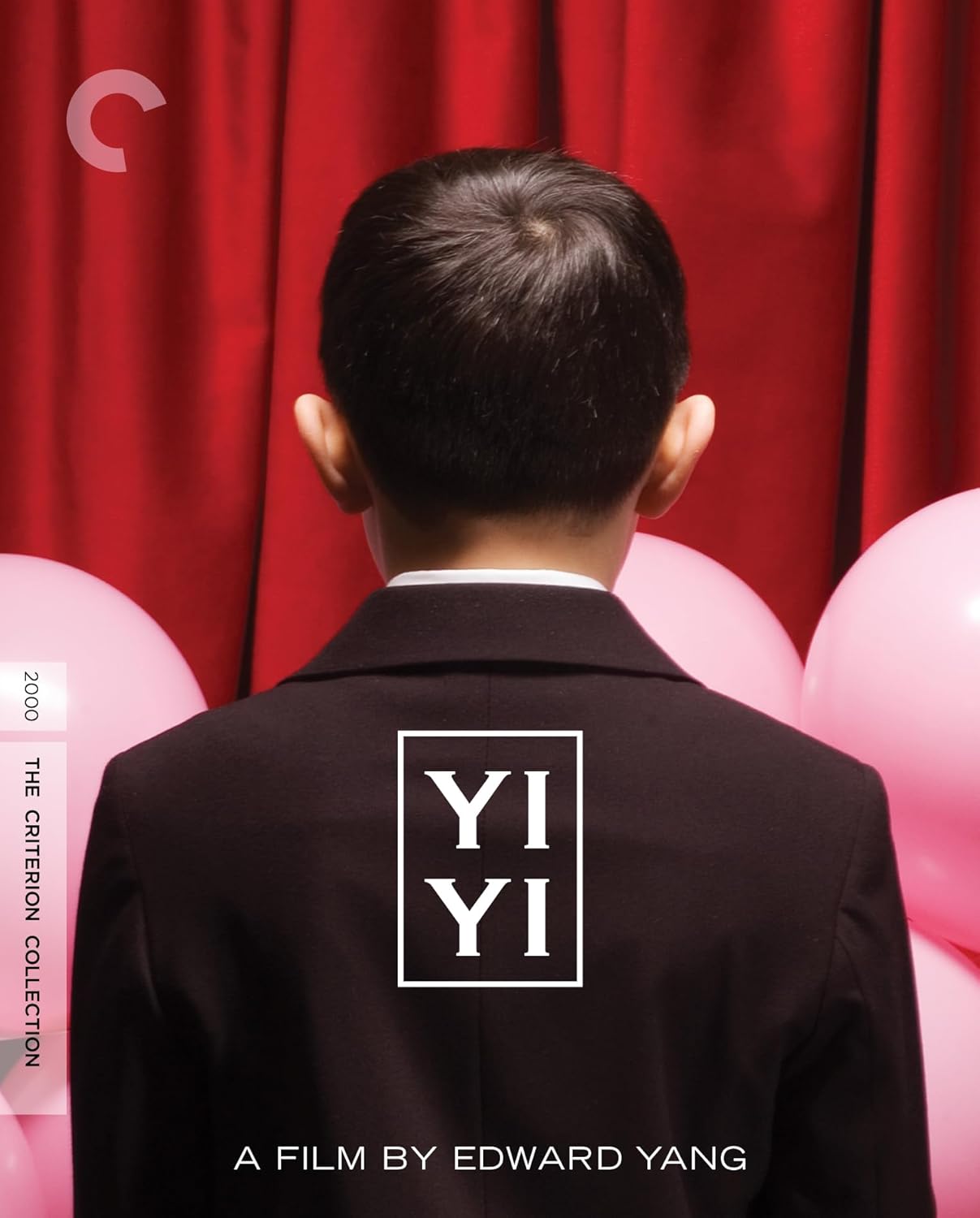 A young boy in a suit faces away from the camera against red curtains.