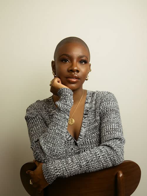 Black woman with shaved head, wearing a patterned top and gold jewelry.