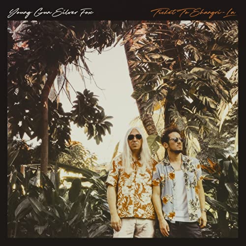 Two men with long hair stand amongst lush tropical foliage.
