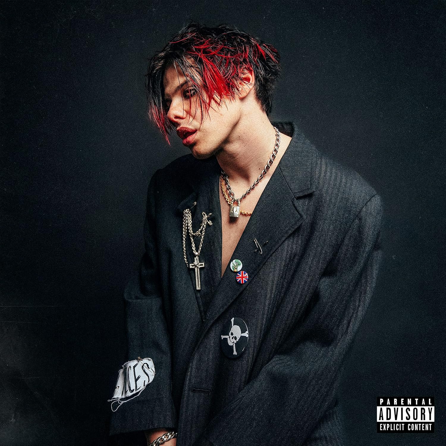 Yungblud with red-streaked hair wearing a dark blazer with pins and chains.