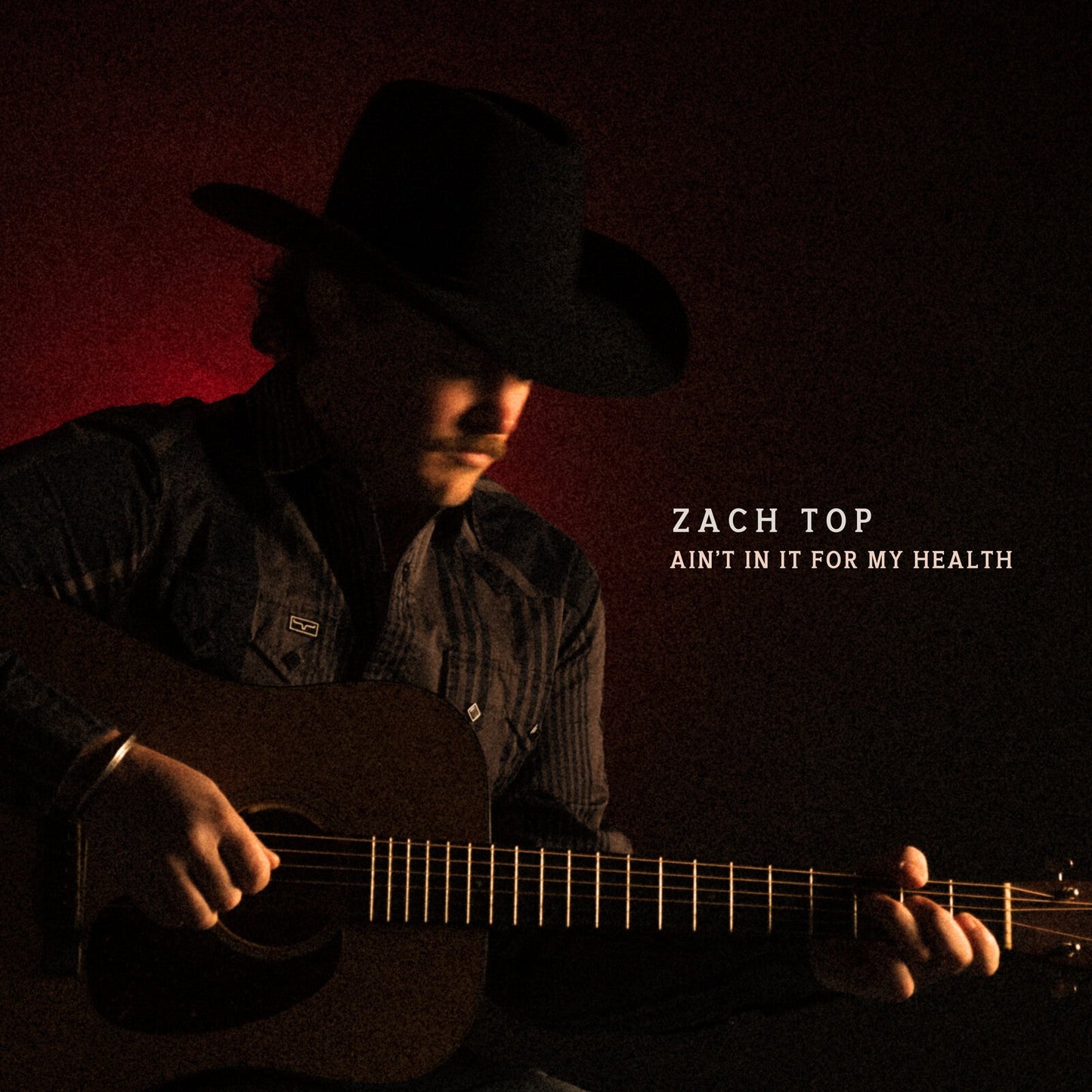 A man in a cowboy hat plays an acoustic guitar against a dark red background.