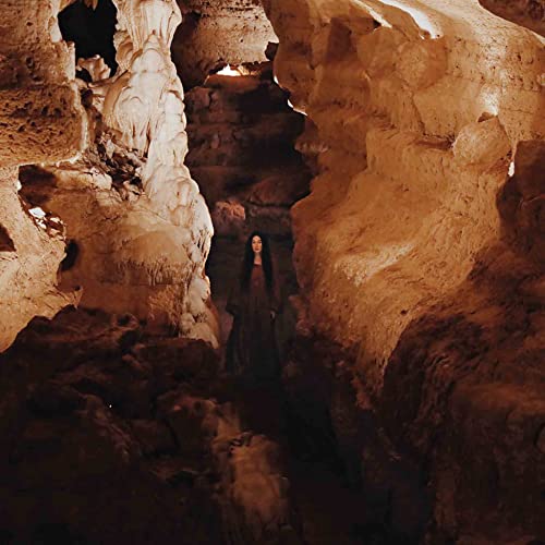 A person with long dark hair stands within a dimly lit, textured rock cave.
