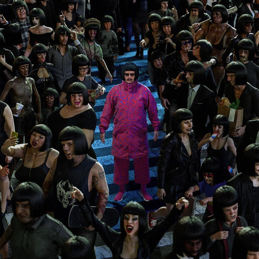 "Man in red stands on stairs surrounded by crowd with identical black bowl haircuts."