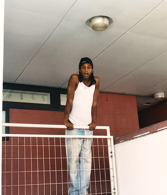 Man in white tank top and black beanie stands behind a white railing.