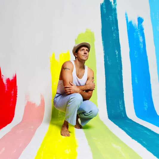 Man crouching against a white wall with colorful brushstrokes, wearing a hat and tank top.