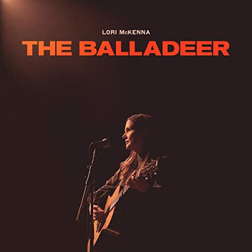 "Woman sings into a microphone, holding a guitar, on a dark background."