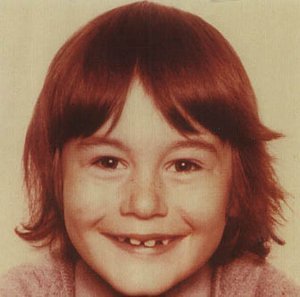 A young child with red hair smiles, showing missing front teeth, against a beige background.