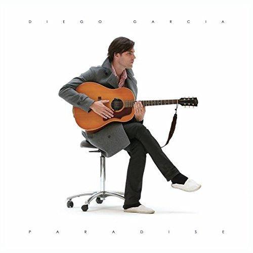 Man sitting on a rolling chair, playing a brown acoustic guitar, against a white background.