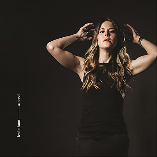 A woman in a black tank top stands against a dark background, hands in her hair.