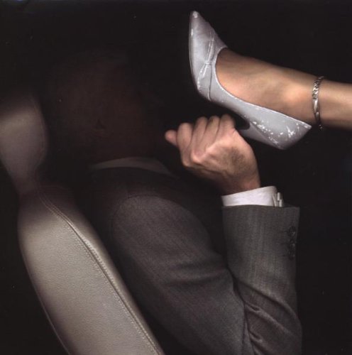A man in a suit holds a woman's silver high-heeled shoe in a dark setting.