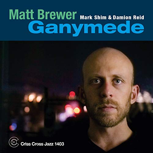 The album cover for "Ganymede" by Matt Brewer features a man with a shaved head and a short beard, wearing a black shirt. The background is dark, with blurred red and white lights visible behind him. The title "Ganymede" is prominently displayed in large blue letters at the top of the image, while the artist's name, "Matt Brewer," is written in green text above it. The names "Mark Shim & Damion Reid" are listed in smaller white