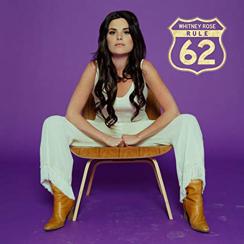 Woman with long dark hair sits on wooden chair against purple background with "Rule 62" sign.